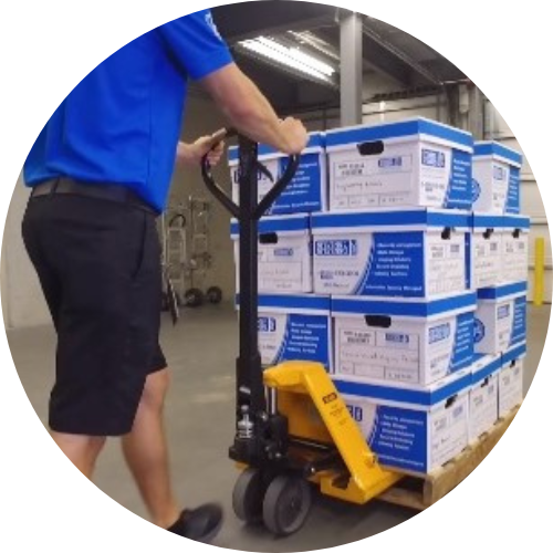 Person in blue uniform shirt and black shorts is using a hydraulic hand truck to move a pallet of labelled archive boxes in a warehouse.