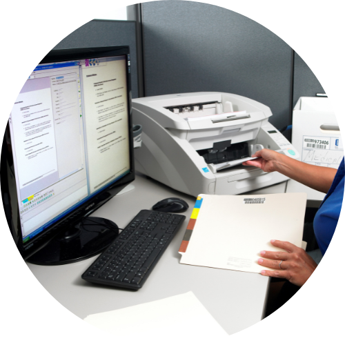 A person sitting at a desk in front of a monitor, holding a file folder and feeding a stack of papers into a scanner